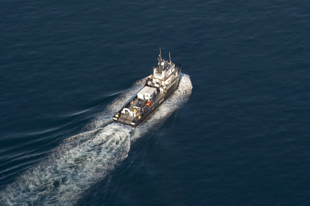A research vessel sails across Monterey Bay. The research vessel has a black hull and white upper deck. Various oceanographic equipment is staged on the ship’s deck. The ship is traveling to the upper right of the frame, with frothy waves in front and leaving a white wake behind. The background is flat, dark-blue ocean.